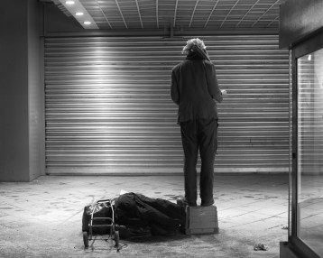 Mann steht auf einer Kiste auf dem Bürgersteig und dirigiert ein unsichtbares Orchester. An Stelle des Orchesters ist ein Roolladen (Rolltor) zu sehen.